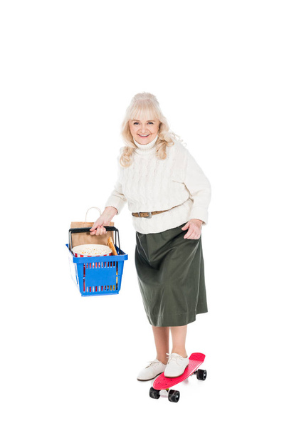 happy senior woman holding shopping basket and riding penny board isolated on white - Foto, Imagem