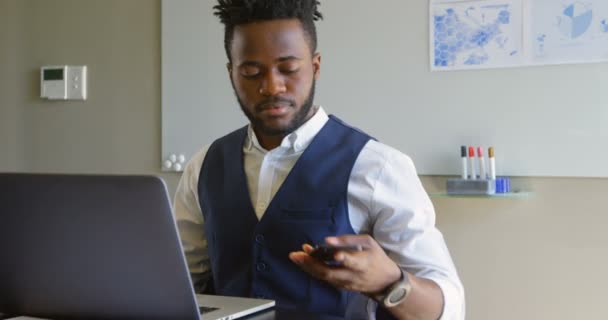 Front view of young black businessman sitting with laptop and talking on mobile phone in office. Businessman working at table in conference room 4k - Séquence, vidéo