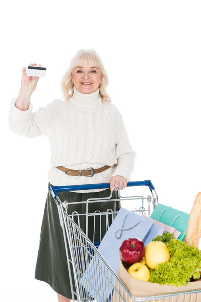 cheerful senior woman holding credit card near shopping trolley isolated on white - Fotoğraf, Görsel