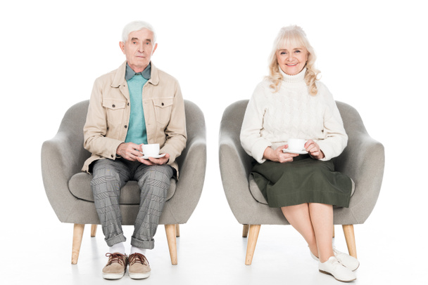 glückliches Rentnerehepaar, das Tassen mit Tee hält, während es in Sesseln isoliert auf weißem Grund sitzt - Foto, Bild