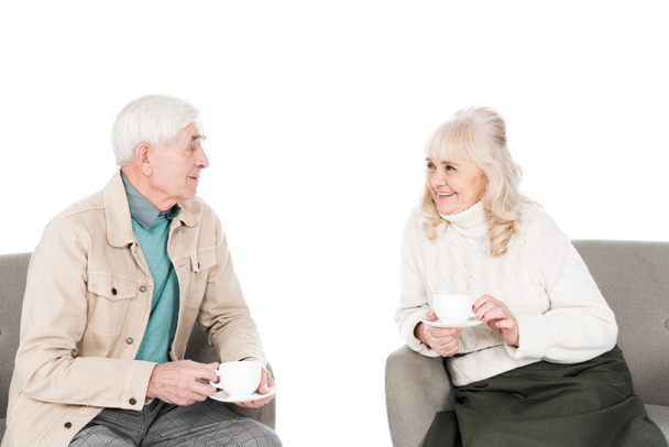 vrolijk gepensioneerde man en vrouw houden cups met thee tijdens de vergadering in Fauteuils geïsoleerd op wit - Foto, afbeelding