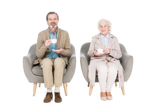 happy senior couple holding cups with tea while sitting in armchairs isolated on white - Photo, image