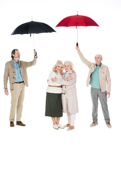 senior men holding umbrellas near women standing and hugging isolated on white - Foto, afbeelding