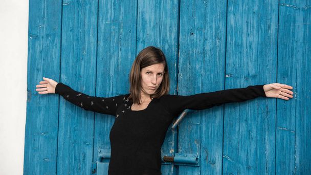 Молода жінка, спираючись на сині сільські дерев'яні двері з широко відкритими руками робить смішне флірт обличчям до камери
. - Фото, зображення