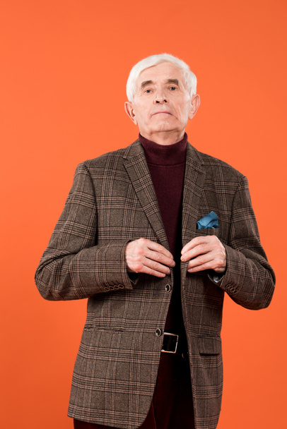 trendy senior man touching jacket while standing isolated on orange  - Zdjęcie, obraz