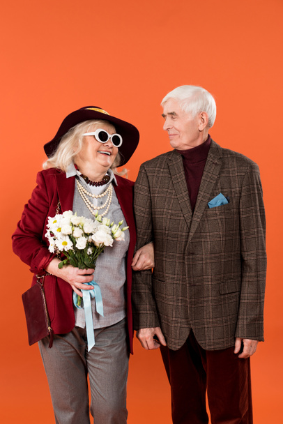 cheerful senior woman in sunglasses and hat looking at husband while holding flowers isolated on orange  - Photo, Image