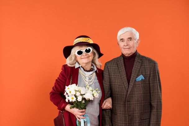 Stilvolle Seniorin mit Sonnenbrille und Hut, die mit ihrem Mann steht und Blumen in Orange hält  - Foto, Bild