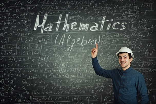 Ein fröhlich positiv denkender junger Ingenieur mit Schutzhelm zeigt mit erhobenem Zeigefinger mit Kreide geschriebene mathematische Formeln und Gleichungen über die Tafel. Bildungskonzept. - Foto, Bild