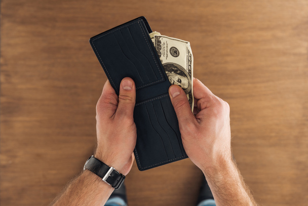 top view of man puting dollars banknotes in wallet on wooden background - Φωτογραφία, εικόνα