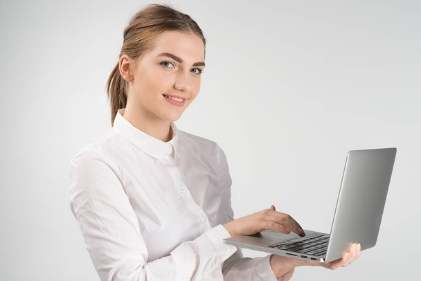Femme d'affaires souriante en chemise blanche tenant un ordinateur portable et tout en se tenant debout et en regardant la caméra
 - Photo, image