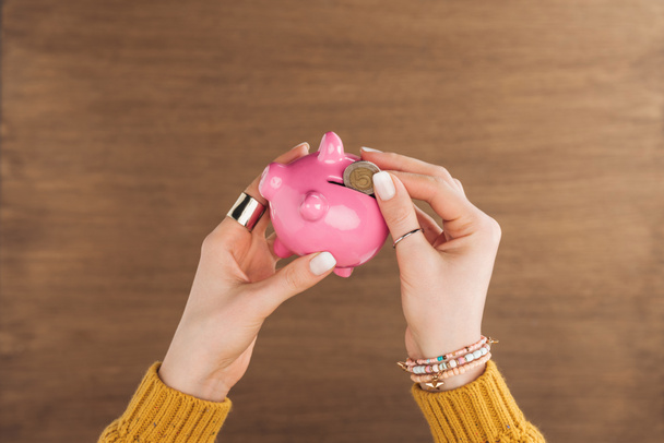 cropped view of woman puting in piggy bank coin on wooden background - Photo, Image