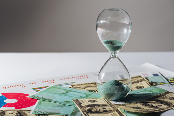 hourglass and coins staks on dollar and euro banknotes with grey background - 写真・画像