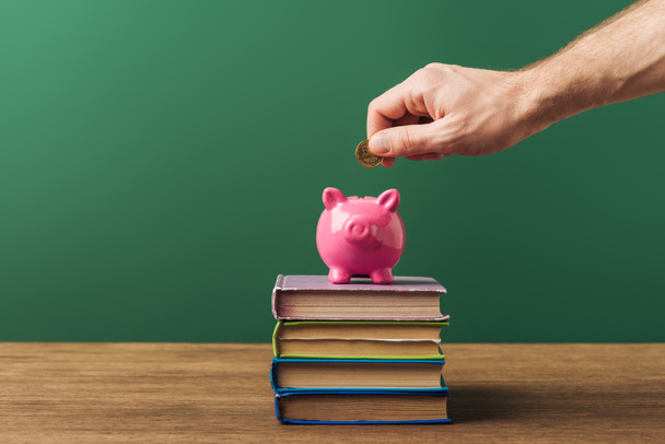 man puting in piggy bank coin on books and wooden table with green background - Foto, afbeelding