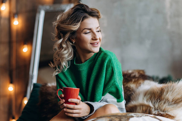 Closeup portrait of a smiling blonde woman in a green sweater with a red cup in her hands. Blurred Background - Foto, Bild