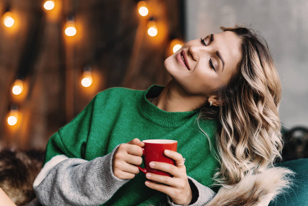 Portrait of a young  blonde woman with closed eyes in a green sweater with a red cup in her hands. Blurred Background - Photo, Image