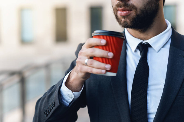 Lähikuva nuori liikemies puku pitää kädessä punainen paperi kuppi kahvia ja aikoo juoda sitä aikaisin aamulla lähellä toimistoa. offee tauko, hämärtynyt tausta
. - Valokuva, kuva
