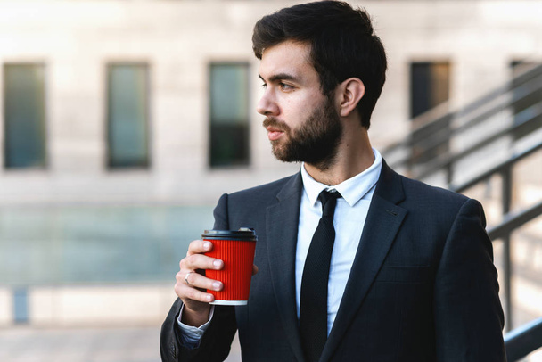 Closeup young businessman in suit holds in hand a red paper cup of coffee and is going to drink it early in the morning near the office. offee break, blurred background. - Fotó, kép