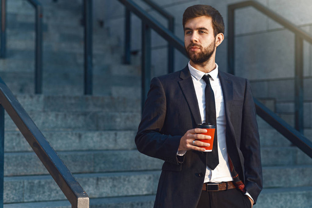 Closeup young businessman in suit holds in hand a red paper cup of coffee and is going to drink it early in the morning near the office. offee break, blurred background. - Photo, Image