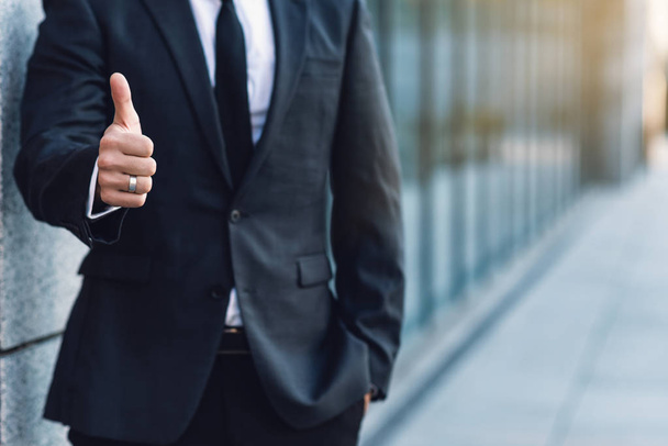 Бизнесмен показывает большой палец вверх, позируя на фоне офисного здания, размытый фон
. - Фото, изображение