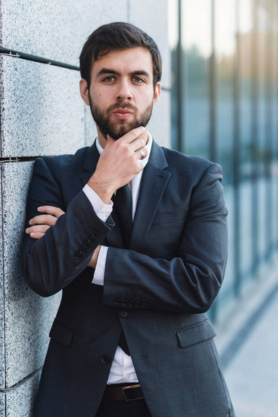 Cloce-up portrait of young businessman holding a beard in a suit posing on background of office building leaning against the wall - Photo, Image