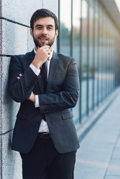 Portrait of young businessman holding a beard in a suit posing on background of office building leaning against the wall - Foto, Bild
