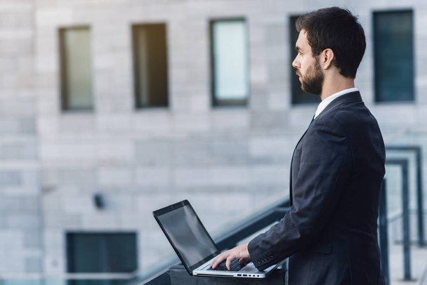 Empresário ao ar livre no fundo de um edifício de negócios trabalhando em um laptop e olhando em linha reta
 - Foto, Imagem
