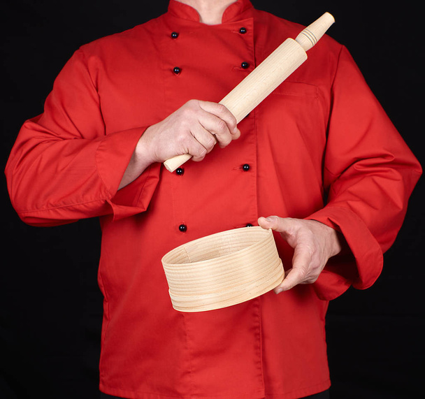 koken in een rood uniform met een houten deegroller en ronde zeef, zwarte achtergrond - Foto, afbeelding