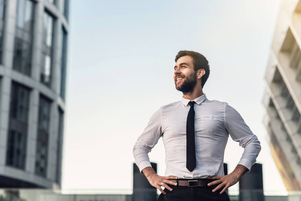 Uomo d'affari in camicia bianca e cravatta sorride felicemente sullo sfondo della città in una giornata di sole tenendo le mani in vita. Guardando lateralmente
 - Foto, immagini