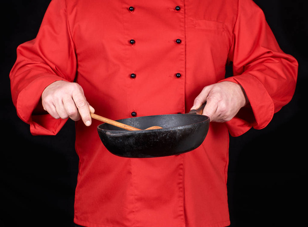 koken in rood uniform holding een lege zwarte koekenpan en een houten lepel, zwarte achtergrond - Foto, afbeelding