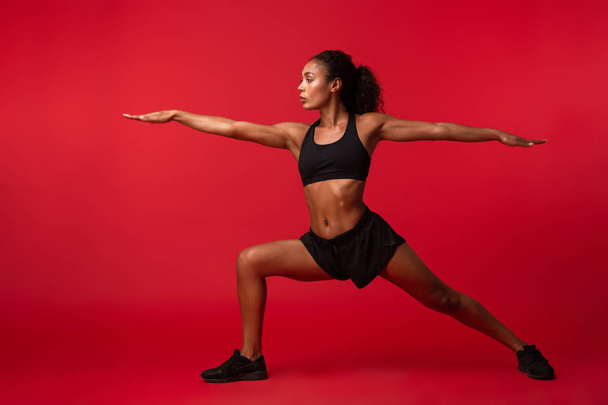 Image of athletic african american woman in black sportswear stretching her body isolated over red background - Fotó, kép