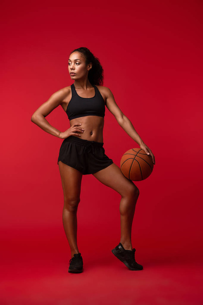 Image of a strong young african sports fitness woman basketball player posing isolated over red wall background holding ball. - Foto, immagini