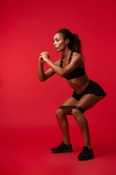 Image of a strong young african sports fitness woman isolated over red wall background make exercises with elastic band. - Valokuva, kuva