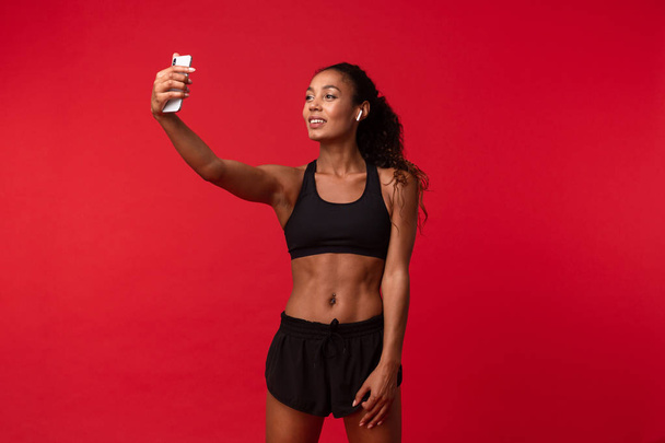 Image of a beautiful young african sports fitness woman posing isolated over red wall background listening music with earphones take a selfie by mobile phone. - Fotografie, Obrázek