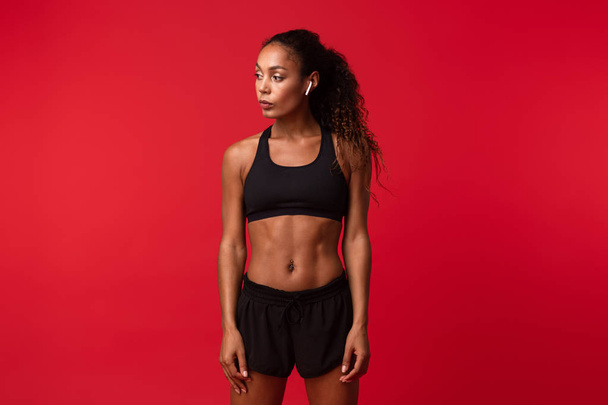 Image of a beautiful young african sports fitness woman posing isolated over red wall background listening music with earphones. - Фото, зображення