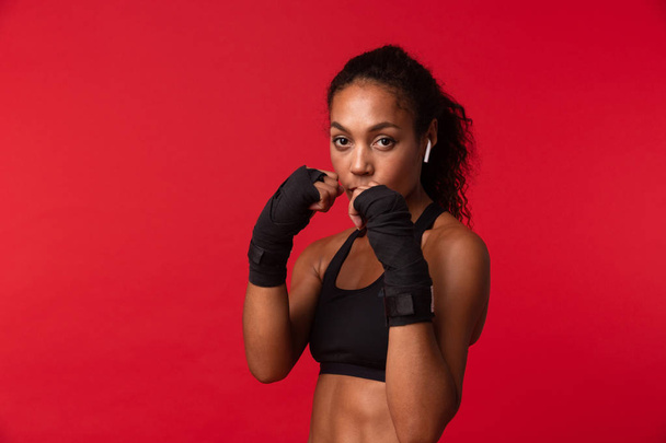 Portrait of strong african american woman in black sportswear boxing with sports bandages on her hands isolated over red background - Foto, imagen