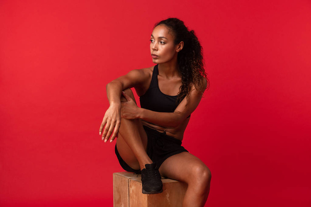 Portrait of beautiful african american woman in black sportswear sitting on box isolated over red background - Фото, зображення