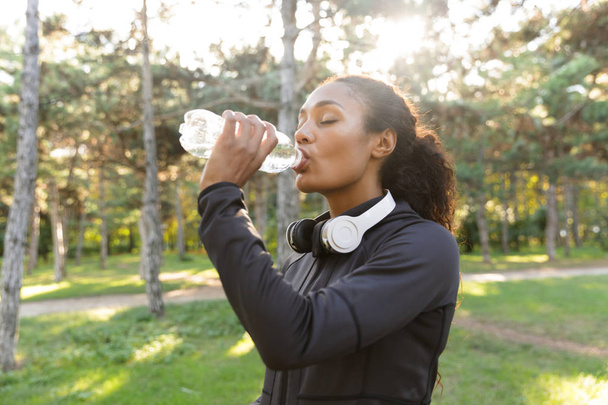 Изображение 20-летней спортсменки в черном спортивном костюме и наушниках, пьющей воду во время прогулки по зеленому парку
 - Фото, изображение