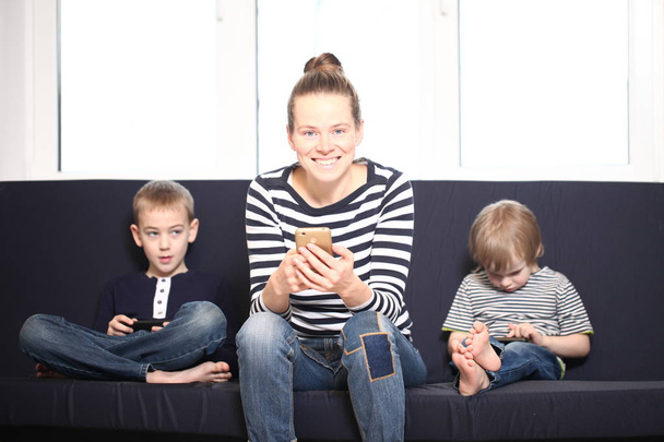 mother with two brothers blond boys on a blue sofa at home playing smartphones - Fotoğraf, Görsel