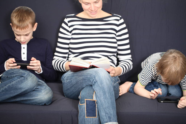 mãe com dois irmãos meninos loiros em um sofá azul em casa jogando smartphones
 - Foto, Imagem