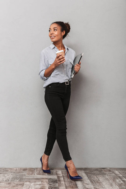 volle Länge einer attraktiven jungen afrikanischen Geschäftsfrau, die Hemd trägt, isoliert über grauen Hintergrund geht, Laptop in der Hand hält, Kaffee zum Mitnehmen trinkt - Foto, Bild