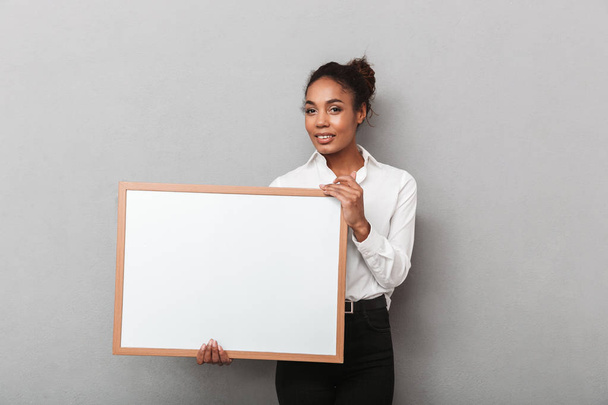 Attractive young african business woman wearing shirt standing isolated over gray background, holding blank board - Valokuva, kuva
