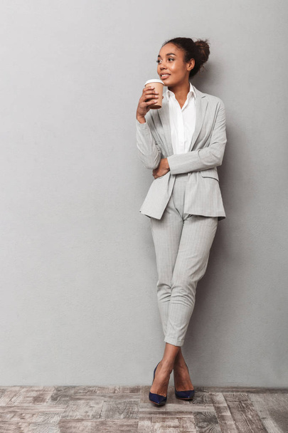 Full length portrait of a confident african business woman wearing suit standing over gray background, holding cup of takeaway coffee - Фото, зображення