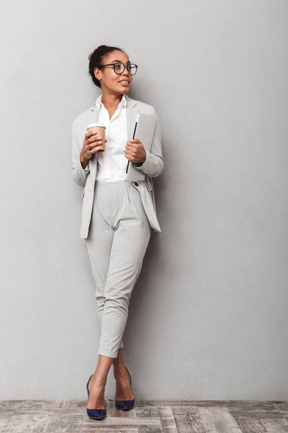 Full length portrait of a confident african business woman wearing suit standing over gray background, holding takeaway coffee, carrying laptop - Fotoğraf, Görsel