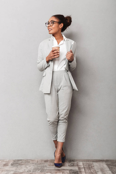 Full length portrait of a confident african business woman wearing suit standing over gray background, holding takeaway coffee, carrying laptop - Fotografie, Obrázek