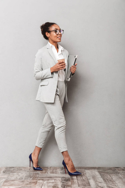 Full length portrait of a confident african business woman wearing suit standing over gray background, holding takeaway coffee, carrying laptop - Фото, изображение