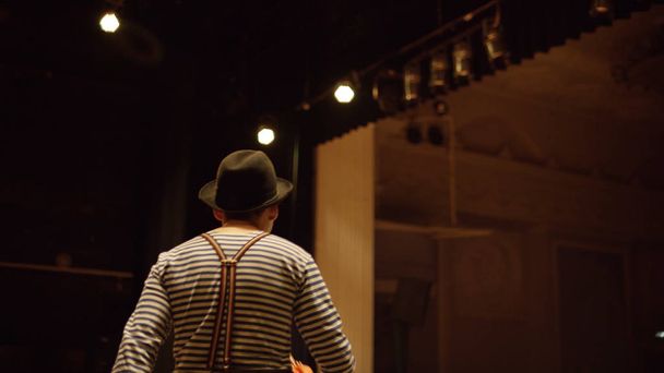 Payaso en el escenario. Vista posterior del payaso con paraguas en las manos con camiseta a rayas y sombrero saliendo de detrás de escena en el escenario
 - Foto, Imagen