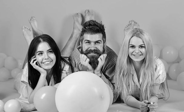 Blonde and brunette on smiling faces have fun with bearded macho. Relations concept. Best friends, lovers near balloons, pink background. Lovers or best friends in pajamas at girlish bedroom party - Фото, изображение