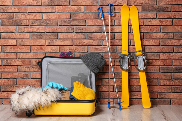 Suitcase with clothes, camera and skis on floor against brick wall. Winter vacation - Foto, Imagem