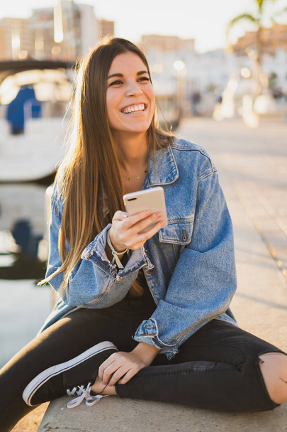 Happy woman smiling and sitting in the sea port using a smartphone and looking at camera - Foto, imagen