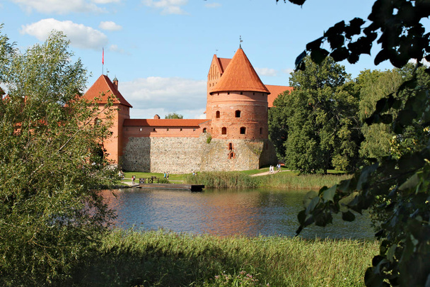 Castelo de Trakai na ilha em Galva Lake. Castel é feito de tijolo vermelho; grama verde ao redor e árvores
 - Foto, Imagem